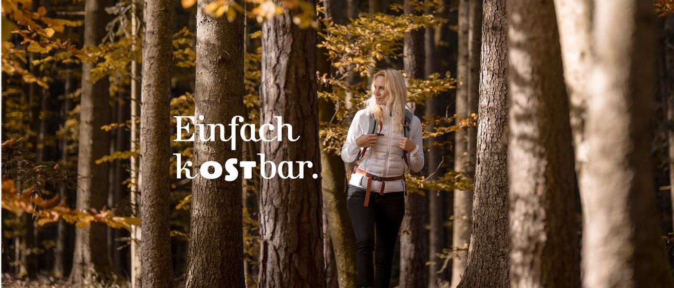 Eine Frau mit blonden Haaren geht in einem Wald mit hohen Bäumen und gefallenen Blättern. Sie trägt eine weiße Jacke, schwarze Hosen und Stiefel. Die Worte 'Einfach kostbar' sind in Weiß auf der linken Seite geschrieben.