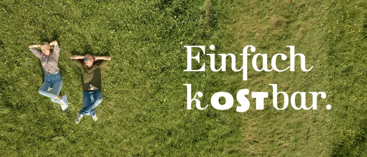 Zwei Personen liegen auf einem Grasfeld, eine trägt eine Sonnenbrille und ein grünes Shirt, die andere ein Blumenhemd und Jeans. Der Satz 'Einfach kostbar' ist prominent in weißem Text dargestellt.