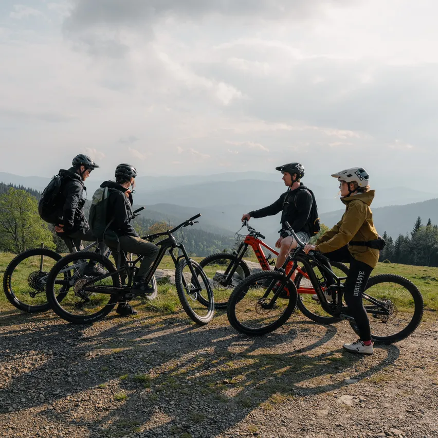 Vier Mountainbiker, alle mit Helmen und Jacken, posieren für ein Foto in den Bergen, mit bewölktem Himmel und fernen Hügeln.