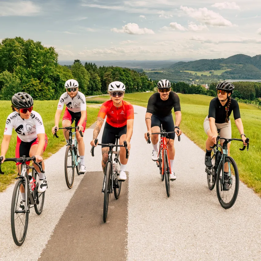 Fünf Radfahrer fahren auf einer kurvenreichen Straße mit einer malerischen Kulisse. Sie tragen Fahrradausrüstung, einschließlich Helmen und Sonnenbrillen. Die Landschaft zeigt üppige Grünflächen und entfernte Berge.