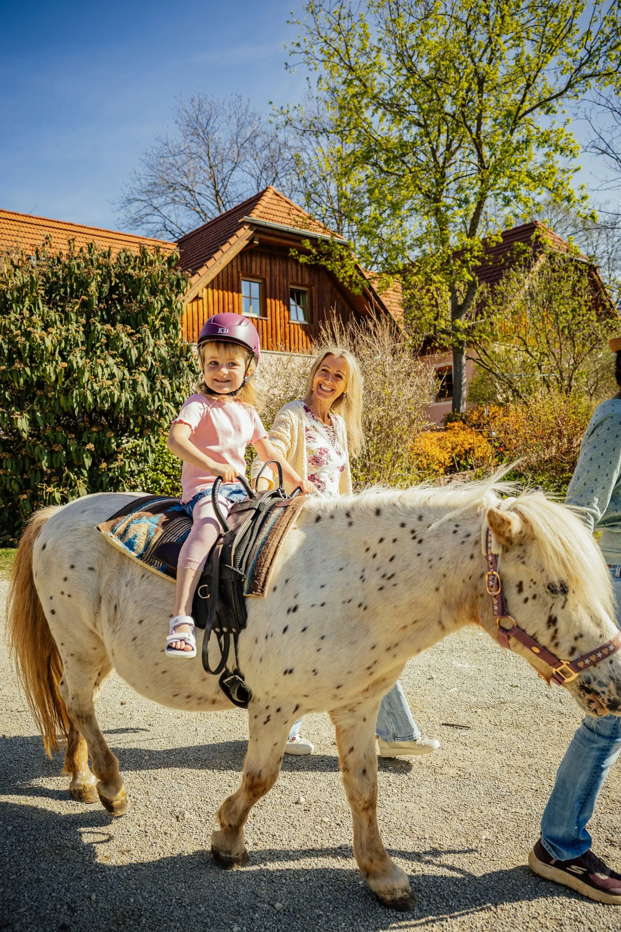 Ein kleines Mädchen reitet auf einem gefleckten Pony, neben ihr geht eine lächelnde Frau. Sie stehen vor einem Haus mit einem roten Dach.