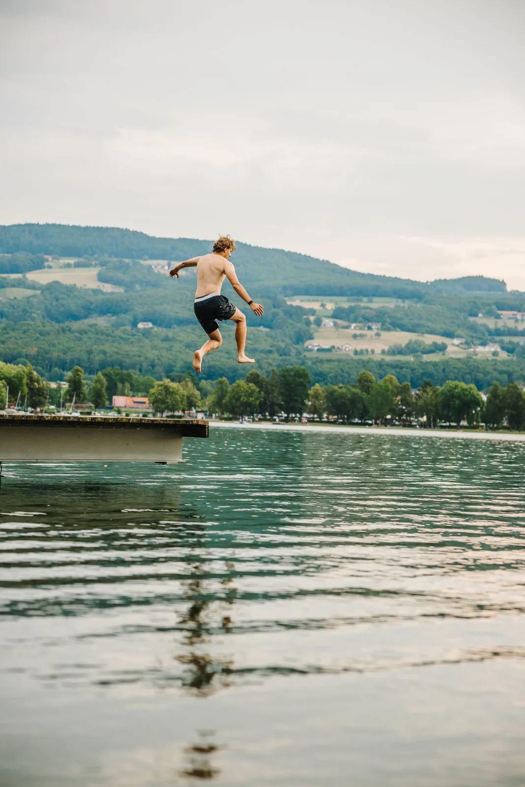 Ein junger Mann springt von einem Steg in einen See, mit Bergen und Bäumen im Hintergrund.