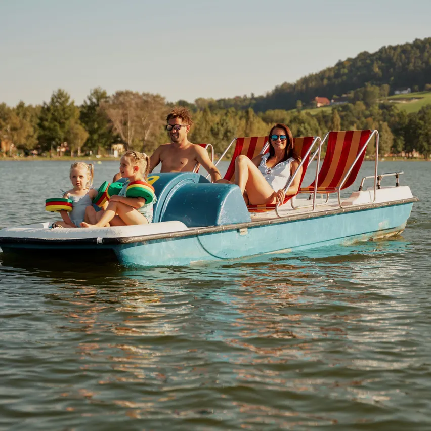 Eine Familie ist auf einem blauen Tretboot mit rot-weiß gestreiften Sitzen. Zwei Kinder und zwei Erwachsene genießen die Fahrt auf einem See mit Bergen im Hintergrund.