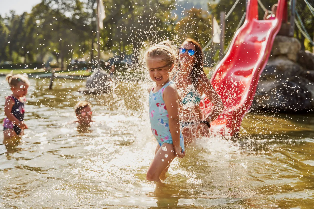 Ein junges Mädchen und eine Frau in Badekleidung spielen in einem Wasserpark, mit einem roten Wasserrutsche im Hintergrund.