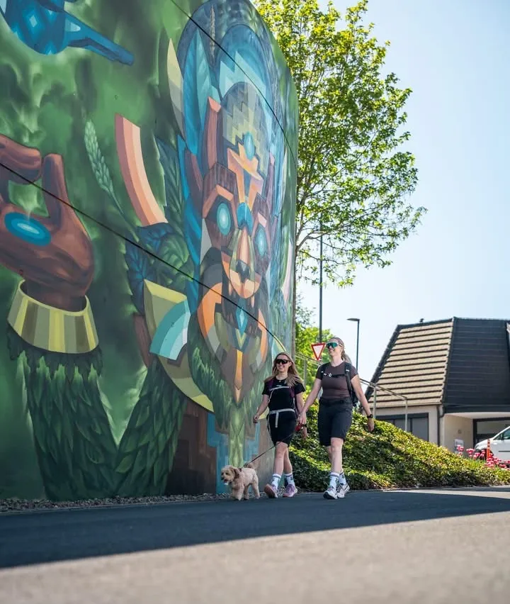 Zwei Frauen spazieren mit ihrem Hund an einer bunten Wandmalerei an einem sonnigen Tag. Ein Haus und ein Verkehrsschild sind sichtbar.