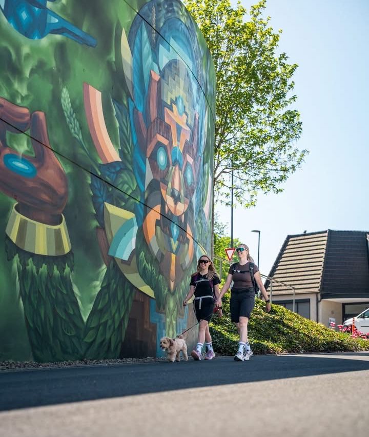 Zwei Frauen spazieren mit ihrem Hund an einer bunten Wandmalerei an einem sonnigen Tag. Ein Haus und ein Verkehrsschild sind sichtbar.