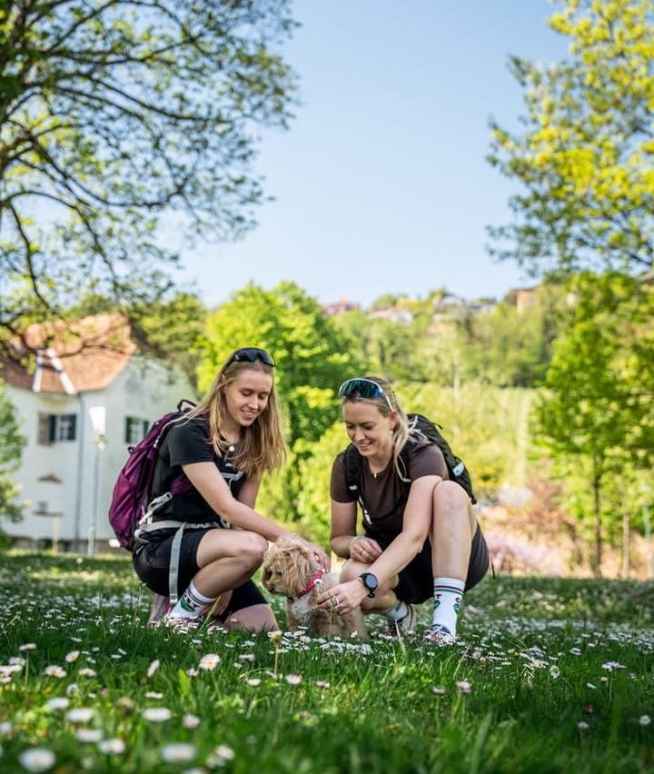 Zwei Frauen in Sportbekleidung mit Rucksäcken knien in einem Feld und streicheln einen kleinen Hund. Im Hintergrund befinden sich Blumen.