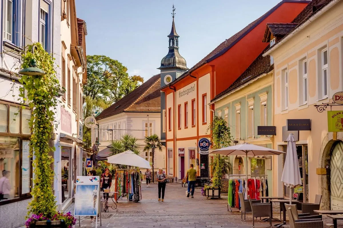 Ein Stadtplatz mit zwei weißen Schirmen. Ein Mann in einem grünen Shirt geht an einem Laden vorbei. Leute stöbern bei Kleidungsständen. Bäume und Gebäude säumen die Straße.