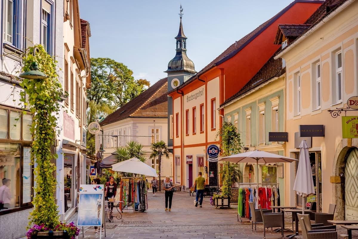 Ein Stadtplatz mit zwei weißen Schirmen. Ein Mann in einem grünen Shirt geht an einem Laden vorbei. Leute stöbern bei Kleidungsständen. Bäume und Gebäude säumen die Straße.