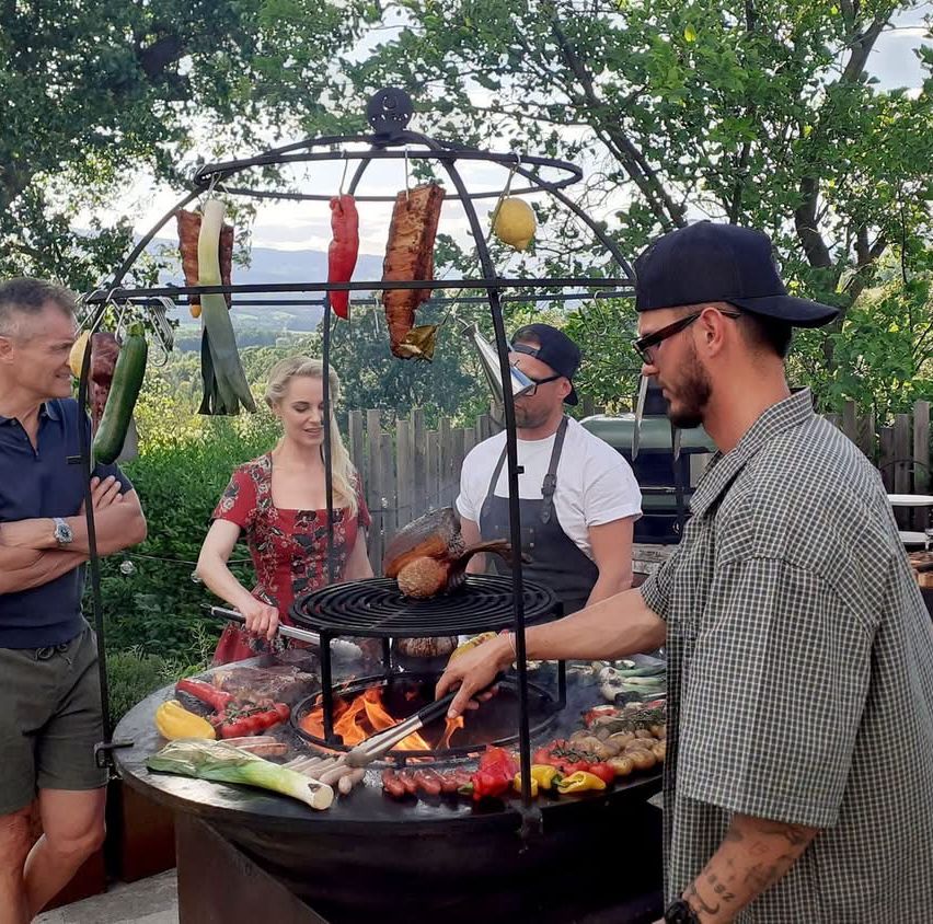 Eine Gruppe von Menschen bereitet ein Grillfest im Freien vor. Eine Frau steht neben dem Grill, während zwei Männer daran arbeiten. Der Grill hat Fleisch, Gemüse und eine über ihm hängende Zitrone.