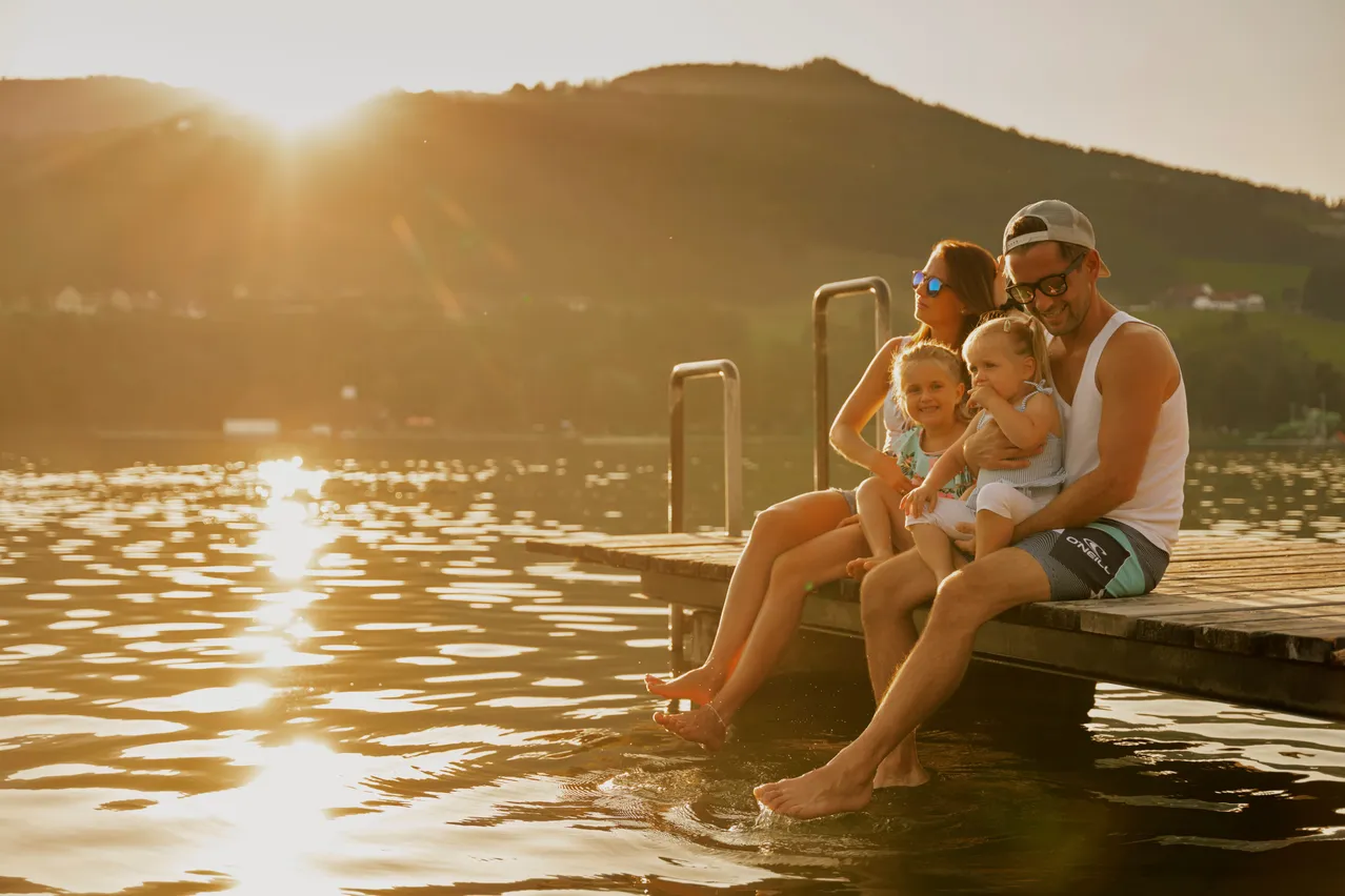 Eine Familie sitzt auf einem Holzsteg mit zwei Kindern und Eltern, Füße im Wasser, genießt den Sonnenuntergang. Dahinter Berge und Wasser mit Reflexionen.