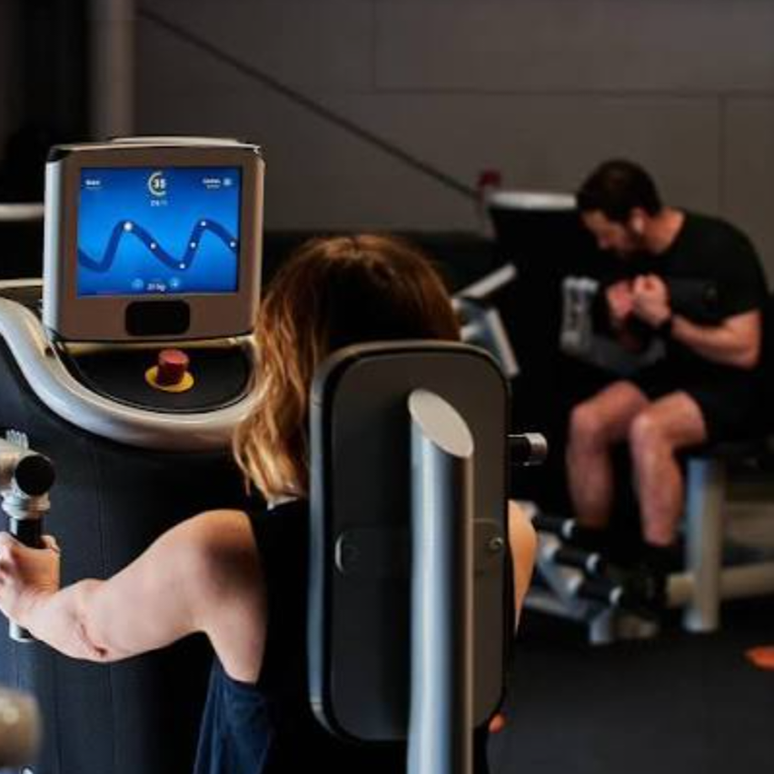 Eine Frau benutzt ein High-Tech-Fitnessgerät, während ein Mann auf einer Bank sitzt, beide tragen schwarze Trainingskleidung.
