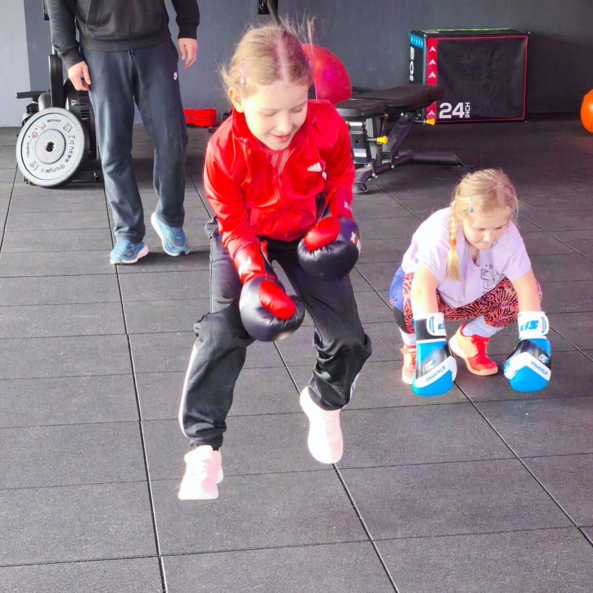Two young girls in boxing gloves practicing in a gym. One girl jumps while the other crouches. A man in sportswear stands behind them.