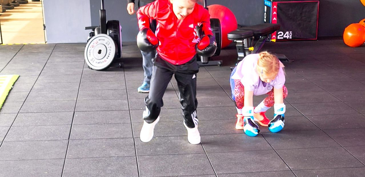 A young boy wearing a red jacket and black boxing gloves is practicing in a gym, while another child crouches nearby. Equipment like a red medicine ball and a black bench press are in the background.