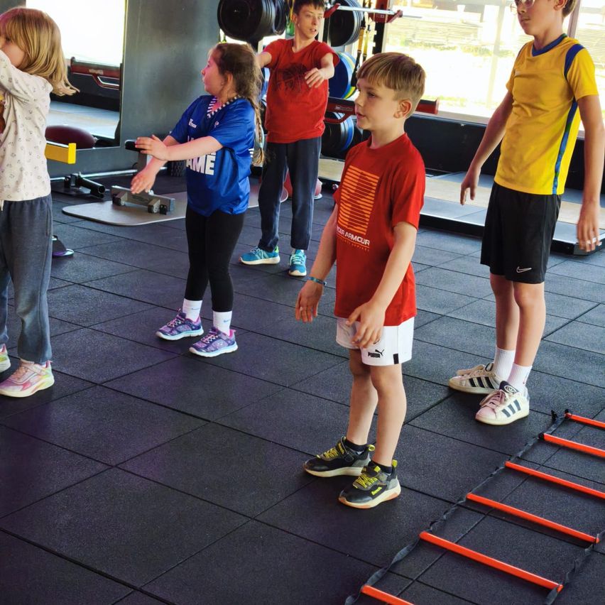 Five children are in a gym, standing on a black floor. One child is stretching and two are facing each other. Equipment and windows are in the background.