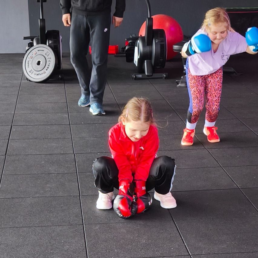 In a gym, two girls wearing boxing gloves practice punching. One crouches on the floor, while another stands nearby with a man. Equipment and a red ball are in the background.