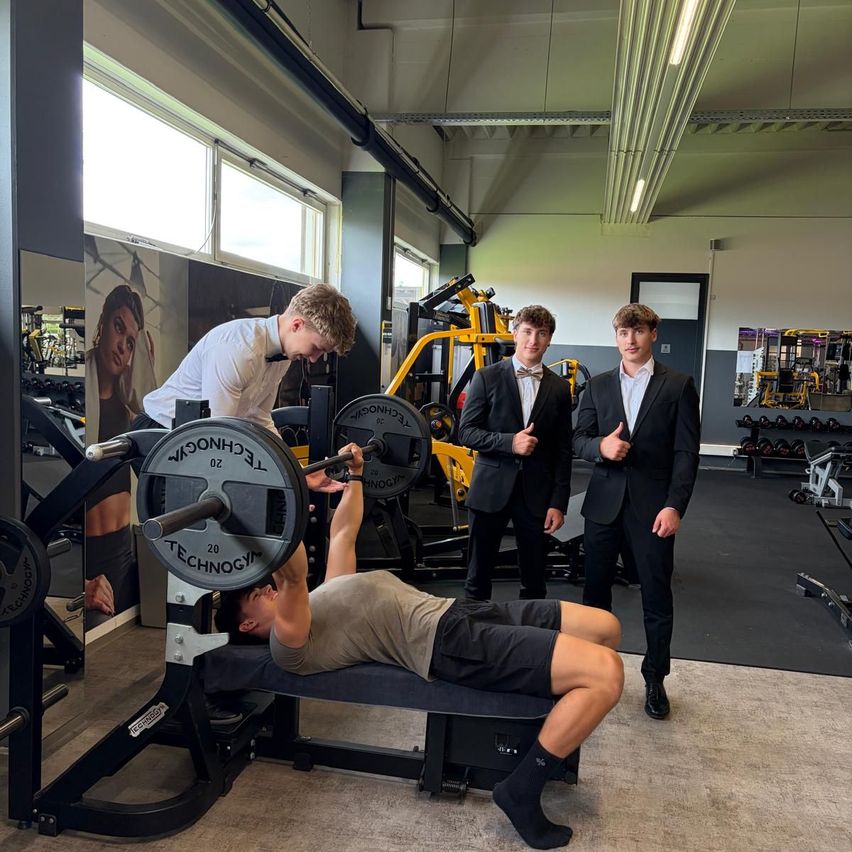 Three young men in suits are in a gym. One is lying on a bench press, another is spotting him, and the third is giving a thumbs up. Various gym equipment is visible in the background.
