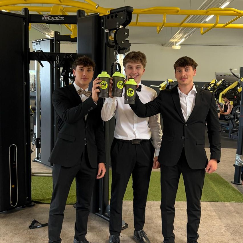 Three young men in formal attire stand in a gym holding green cups with yellow text. One man holds the cups while another man stands behind them in a similar pose. Gym equipment is in the background.