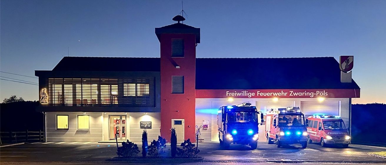 Feuerwehrstation mit mehreren Fahrzeugen draußen bei Nacht. Roter Turm, große Fenster und beleuchtetes Schild mit der Aufschrift 'Freiwillige Feuerwehr Zwaring-Pols'.
