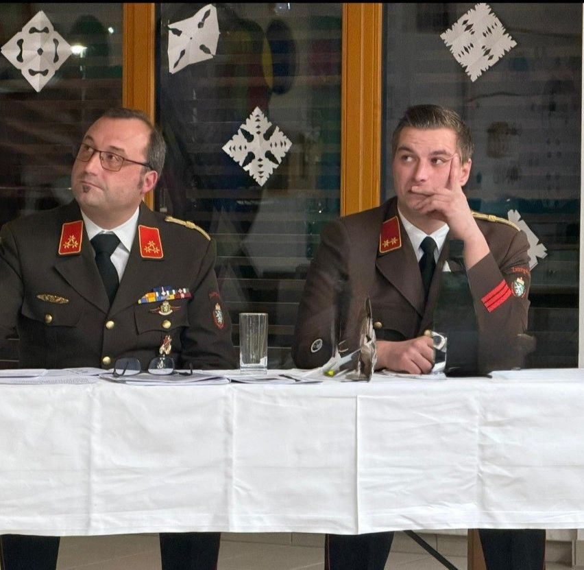 Zwei Militäroffiziere sitzen an einem Tisch mit Gläsern und Papieren vor ihnen. Sie tragen Uniformen mit Aufnähern an den Schultern. Der Mann auf der rechten Seite hat seine Hand im Kinn. Hinter ihnen hängen dekorative Schneeflocken an der Glasscheibe.