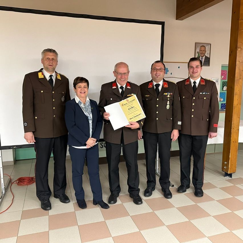 Fünf Personen stehen in einem Raum und lächeln. Eine Frau hält ein Zertifikat. Hinter ihnen befinden sich eine Tafel und ein Bilderrahmen an der Wand.