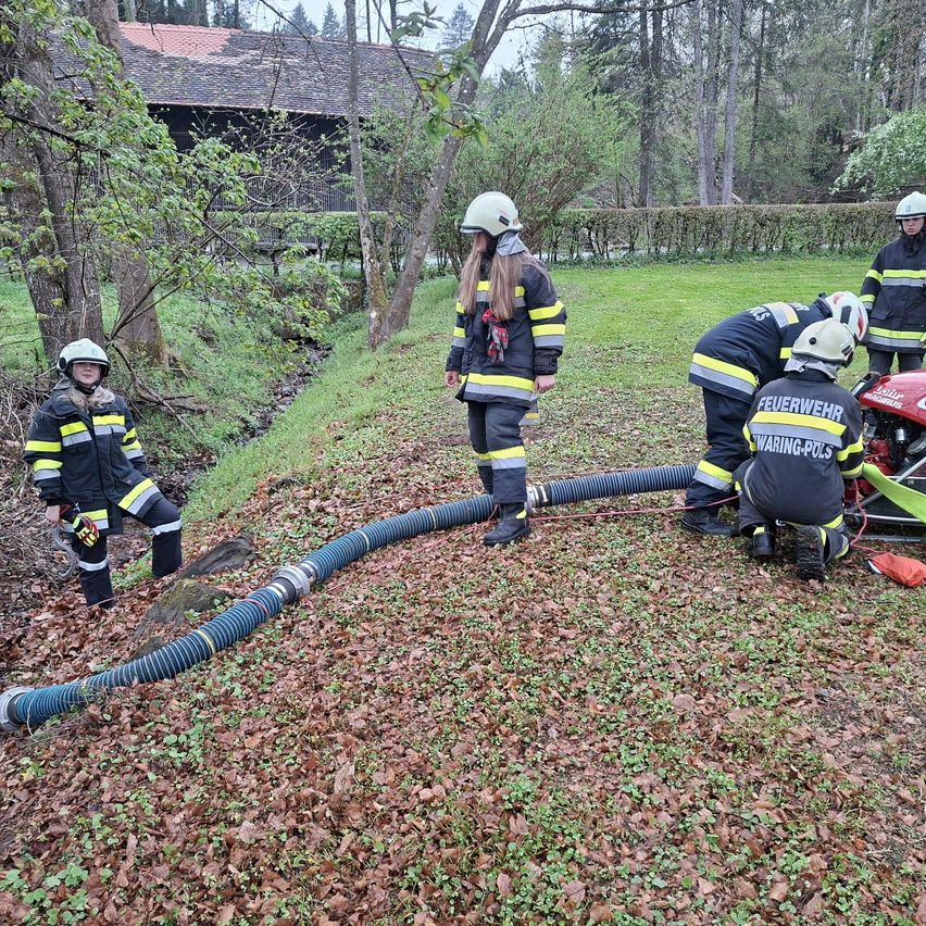 Firefighters in yellow and black uniforms are practicing with a hose on the grassy ground. The hose is laid out on the ground. They are standing near a water stream.