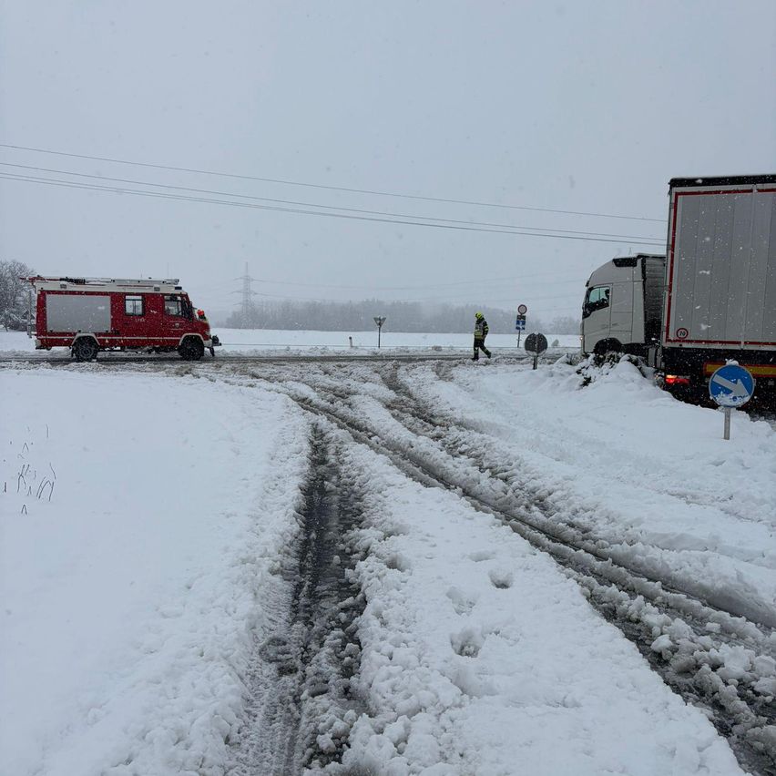 Ein roter Feuerwehrwagen ist auf einer verschneiten Straße geparkt, mit einer Person, die in der Nähe geht. Ein weißer Lkw steht rechts mit einem blauen Schild davor. Schnee bedeckt den Boden, und Strommasten sind in der Ferne.