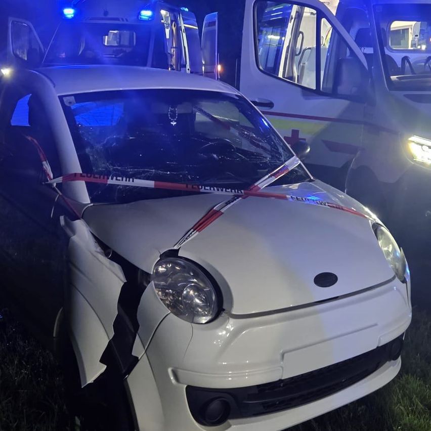 Ein weißer Wagen mit zertrümmerter Front ist mit rot-weißem Band abgesperrt. Dahinter parken zwei Krankenwagen mit eingeschaltetem Blaulicht.