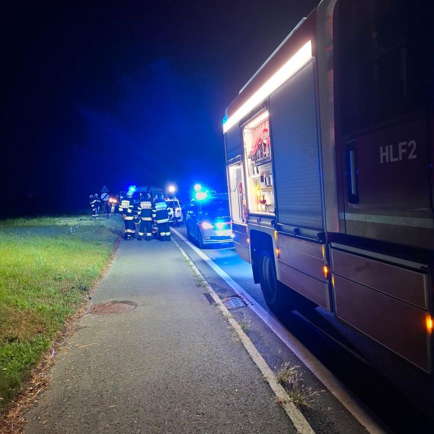 Rettungskräfte in Uniform versammeln sich nachts auf einer Straße, mit einem Feuerwehrwagen und einem Polizeiwagen in der Nähe.