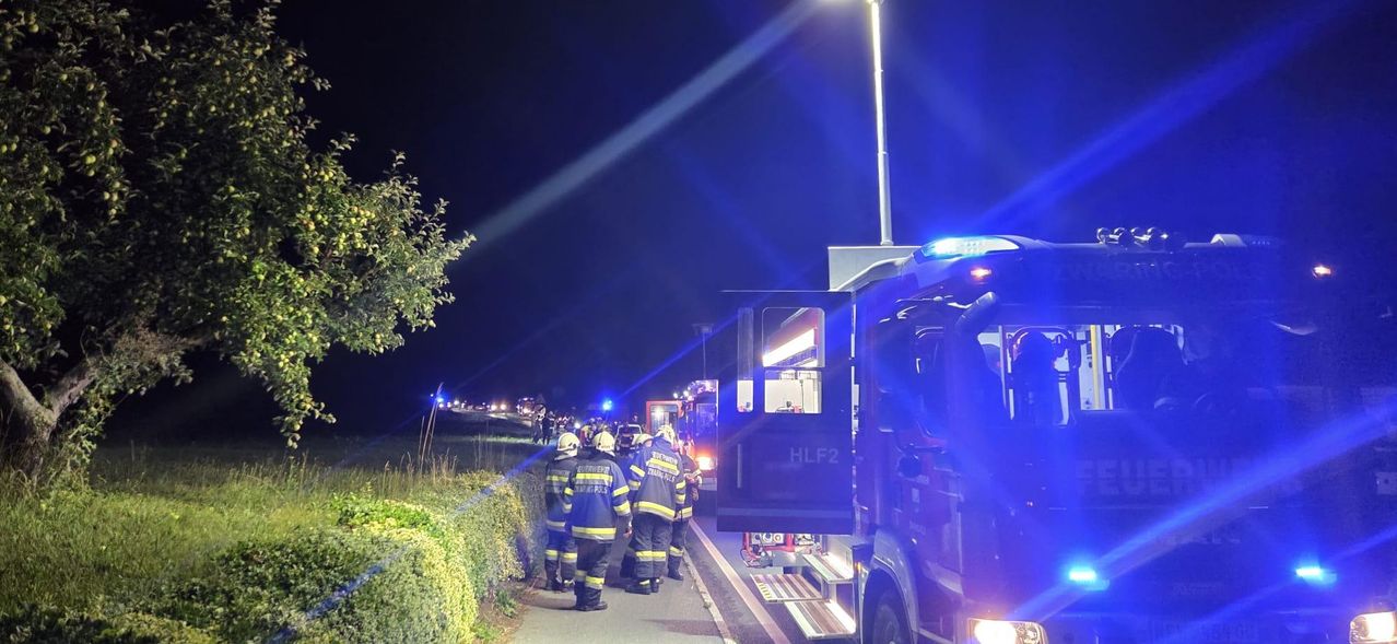 Feuerwehrleute in Uniform stehen nachts am Straßenrand, mit Einsatzfahrzeugen und blinkenden blauen Lichtern.