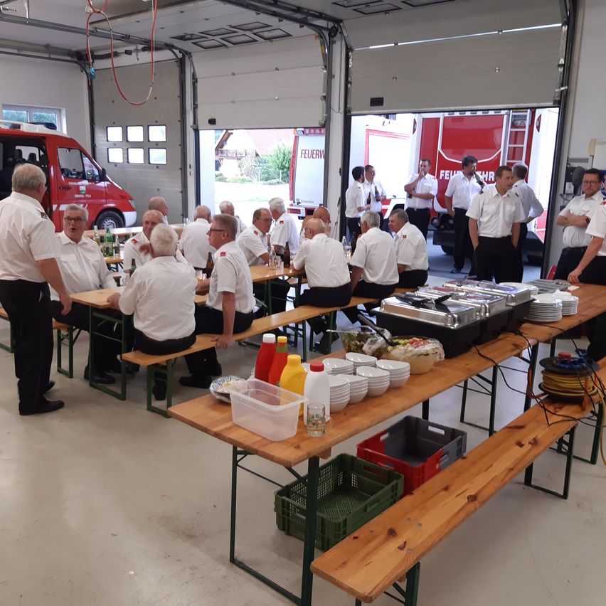 Eine Gruppe von Feuerwehrleuten ist in einer Feuerwache versammelt, sitzt an Tischen mit Essen und Getränken, während andere herumstehen und ein Feuerwehrwagen draußen geparkt ist.