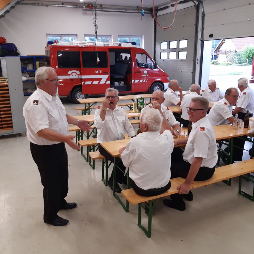 Eine Gruppe älterer Männer in weißen Uniformen sitzt an einem Tisch in einer Garage, mit einem roten Notfallfahrzeug dahinter.