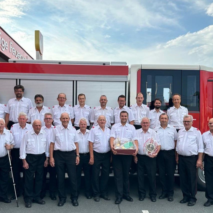 Eine Gruppe von Feuerwehrleuten in weißen Uniformen und schwarzen Hosen posiert für ein Foto vor einem roten Feuerwehrwagen. Einige von ihnen halten Geschenke.