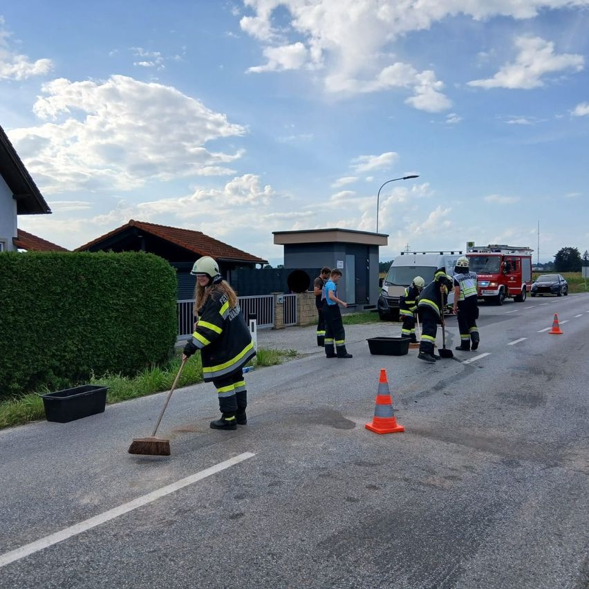 Eine Gruppe von Feuerwehrleuten reinigt eine Straße mit Besen und Kehrschaufeln, wobei Verkehrskegel den Bereich abgrenzen. Der Himmel ist blau mit verstreuten Wolken.
