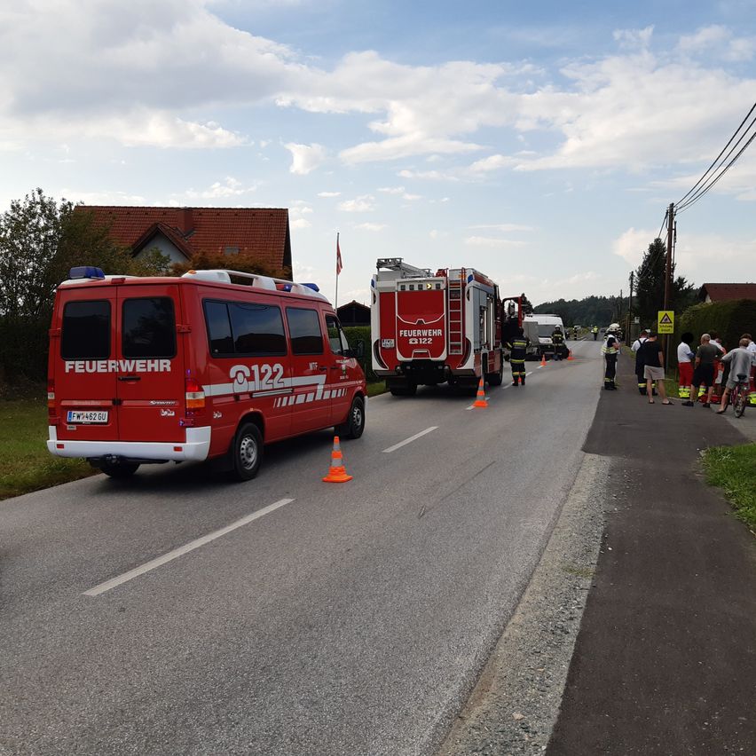 Zwei Feuerwehrfahrzeuge sind am Straßenrand geparkt, mit Menschen, die herumstehen. Es gibt Verkehrskegel und eine Fahne an der Seite.