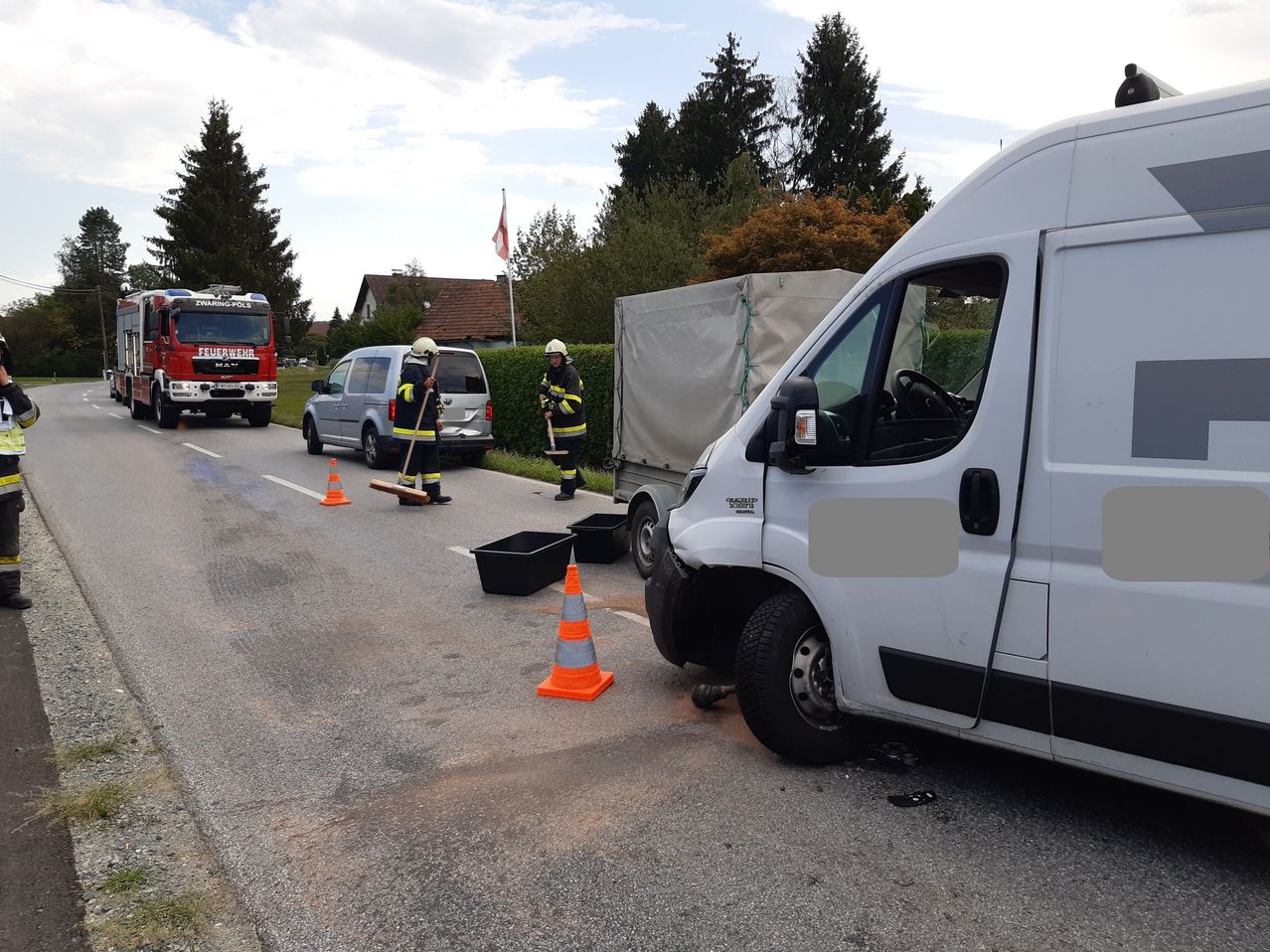 Zwei Feuerwehrleute räumen Trümmer von einem Verkehrsunfall auf, bei dem ein weißer Lieferwagen und ein roter Feuerwehrwagen beteiligt waren. Verkehrskegel und ein silberner Van sind in der Nähe.