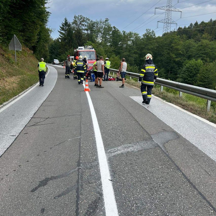 Rettungskräfte kümmern sich auf einer Straße um einen Unfall, wobei ein Feuerwehrwagen in der Nähe geparkt ist und Verkehrskegel den Bereich abgrenzen.