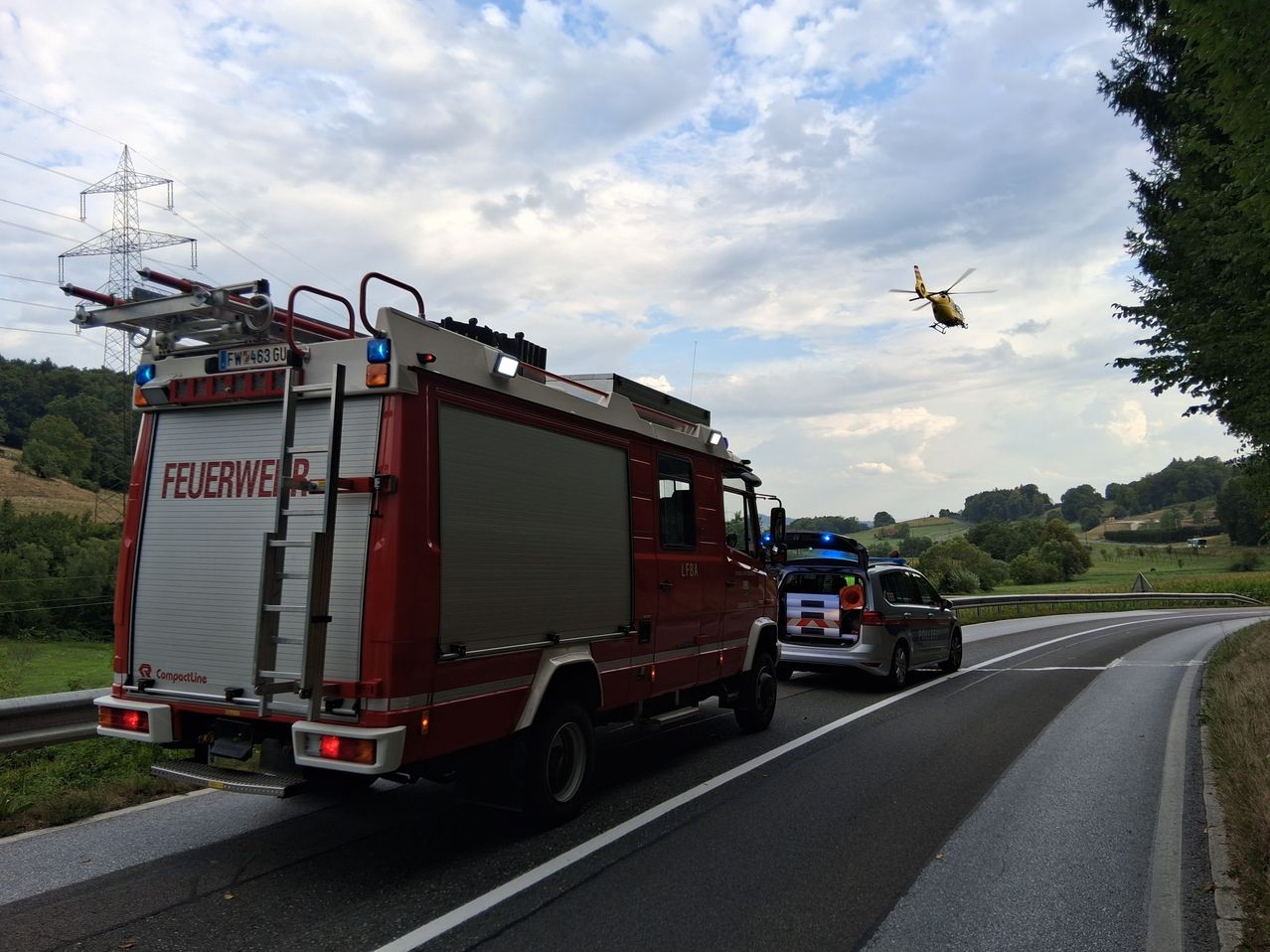 Ein Feuerwehrwagen steht am Straßenrand, ein Hubschrauber fliegt darüber. Ein Auto ist auf der gegenüberliegenden Straßenseite geparkt.