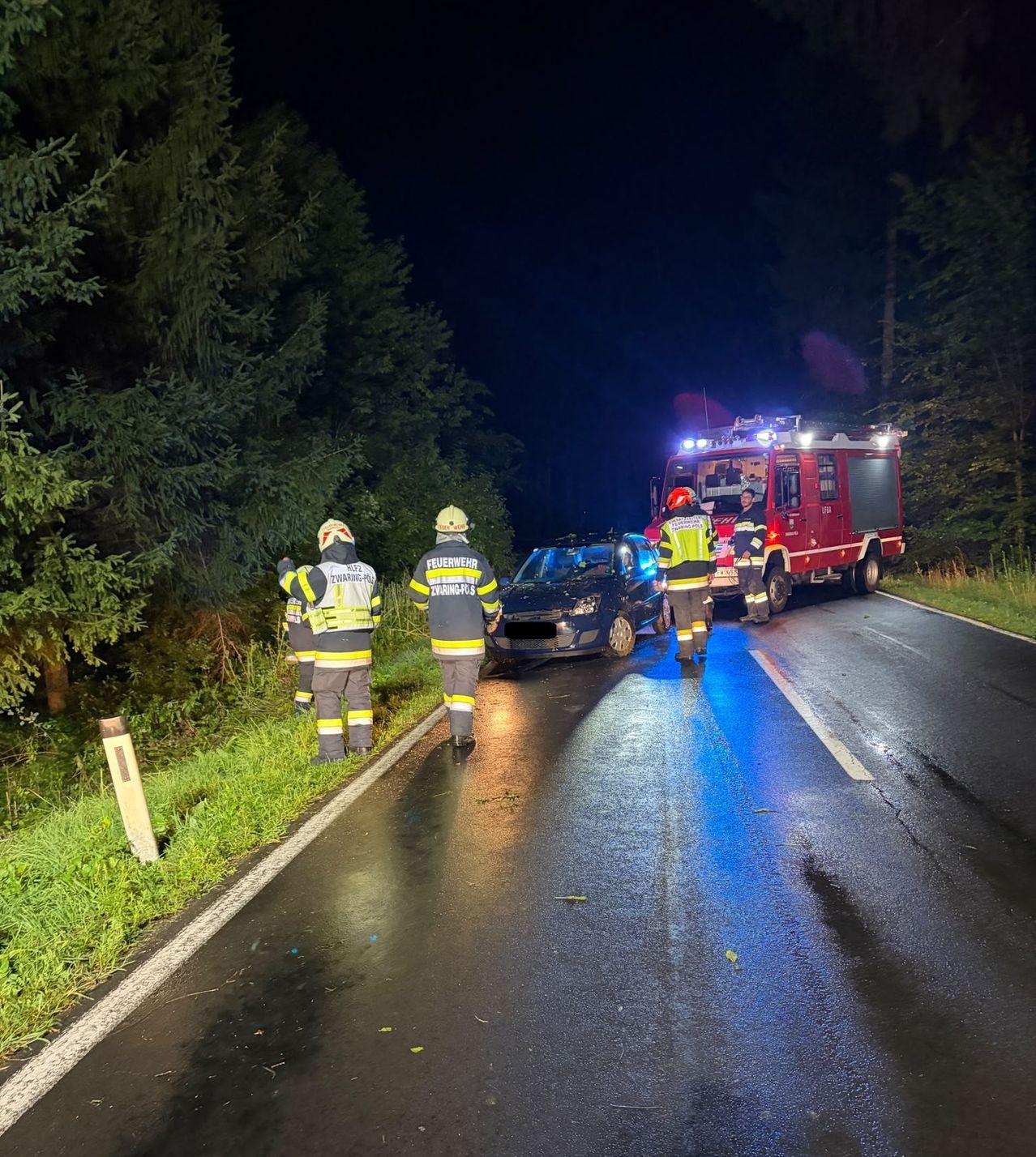 Eine Gruppe von Feuerwehrleuten in Uniform kümmert sich um einen Autounfall auf einer dunklen Straße bei Nacht. Ein roter Feuerwehrwagen mit eingeschalteten Lichtern ist in der Nähe geparkt.