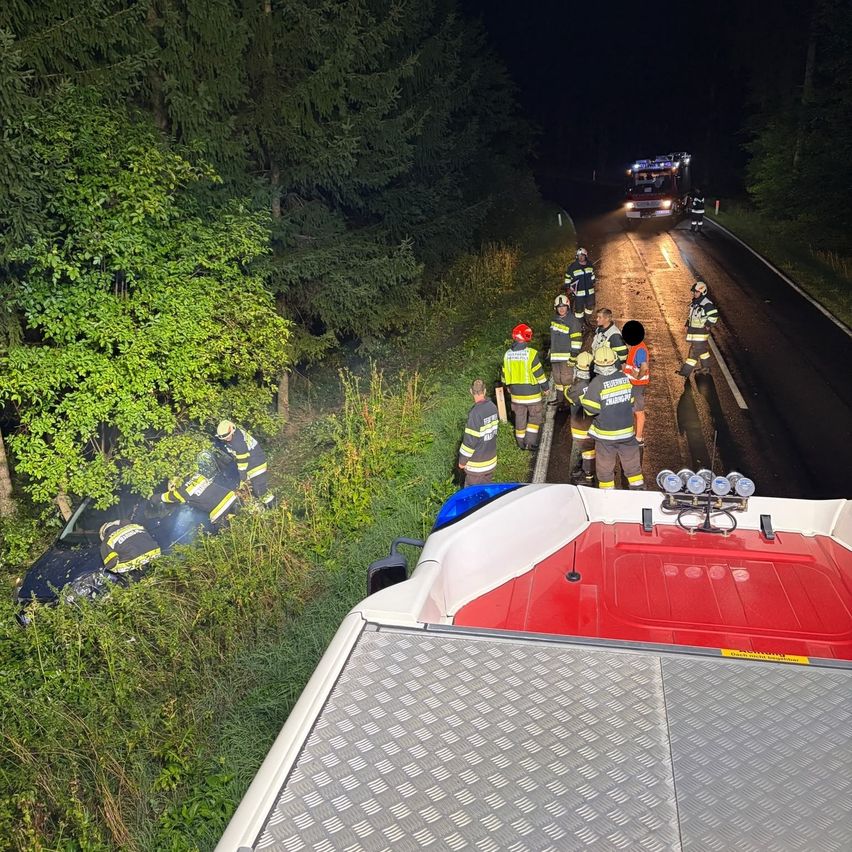 Rettungskräfte kümmern sich um einen Verkehrsunfall auf einer dunklen Straße, wobei ein Feuerwehrwagen in der Ferne zu sehen ist.