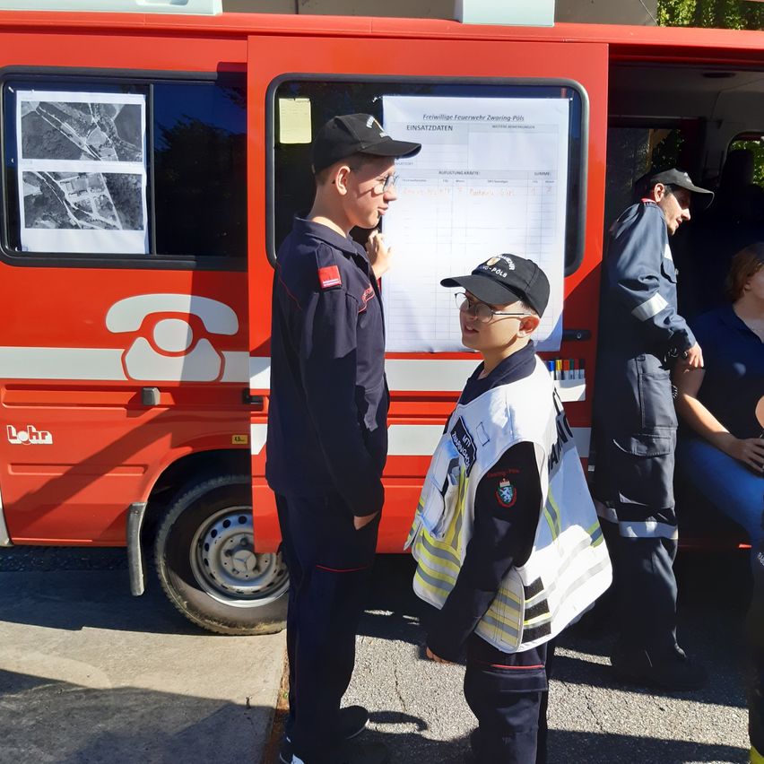 Zwei Feuerwehrleute in Uniform stehen vor einem Feuerwehrauto mit einem Schild. Ein junger Junge in einer Warnweste steht neben ihnen. Eine Frau sitzt im Hintergrund.