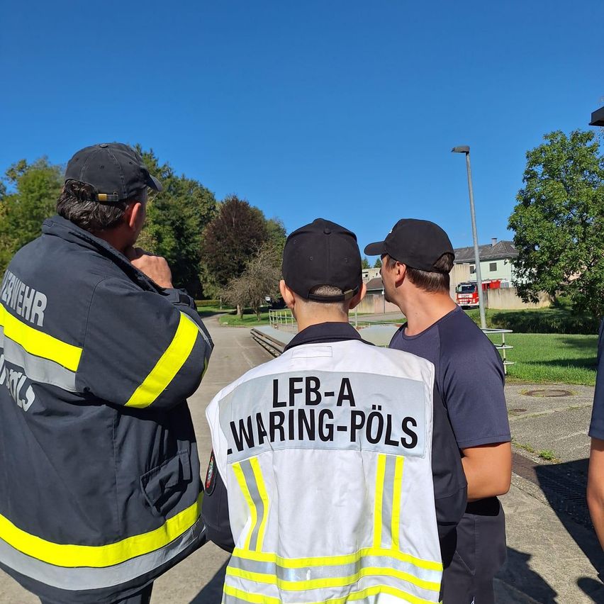 Drei Feuerwehrleute in Warnwesten stehen auf einer Betonplattform, einer trägt einen Hut und ein anderer eine Warnweste mit der Aufschrift 'LFB-A WARING-POLS'.