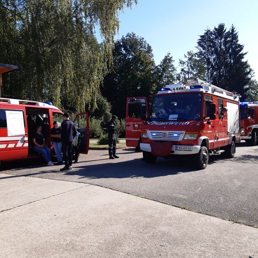 Eine Gruppe von Menschen versammelt sich um einen roten Feuerwehrwagen mit offener Tür. Ein Mann in schwarzer Uniform steht in der Nähe. Ein weiterer Feuerwehrwagen ist dahinter geparkt. Bäume und ein Gebäude sind im Hintergrund zu sehen.