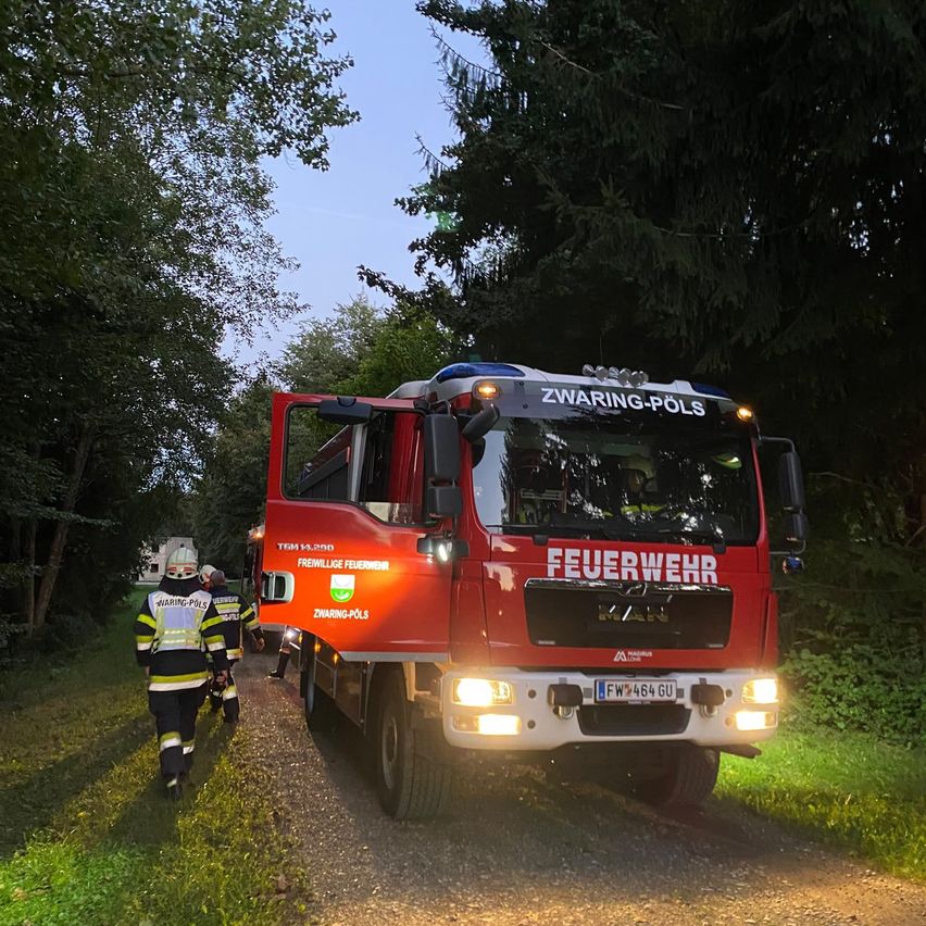 Ein roter Feuerwehrwagen mit offener Tür, geparkt auf einem Schotterweg, mit Feuerwehrleuten in der Nähe.