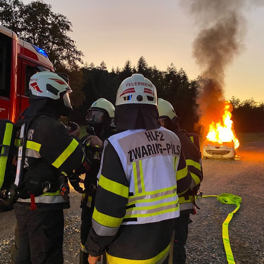 Feuerwehrleute in voller Ausrüstung stehen um ein brennendes Auto herum, einer hält einen Schlauch. Das Auto steht auf einem Schotterweg, und Rauch steigt in den Himmel.
