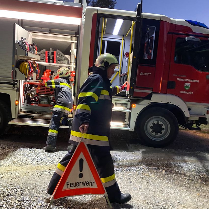 Zwei Feuerwehrleute bereiten sich darauf vor, in einen Feuerwehrwagen einzusteigen, einer hält ein Warnschild. Der Wagen ist geöffnet und zeigt sein Inneres mit verschiedenen Ausrüstungsgegenständen.