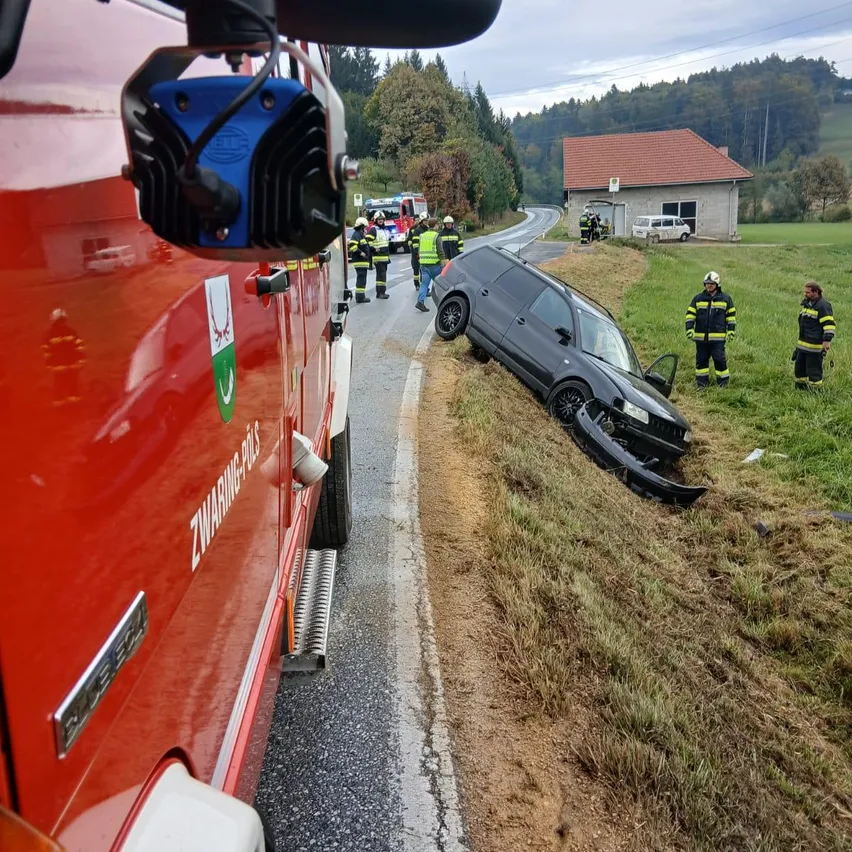 Ein roter Feuerwehrwagen ist am Straßenrand geparkt, mit einem beschädigten schwarzen SUV, der auf der Seite auf dem Straßenrand liegt. Mehrere Feuerwehrleute kümmern sich um die Szene, und ein Haus ist im Hintergrund zu sehen.