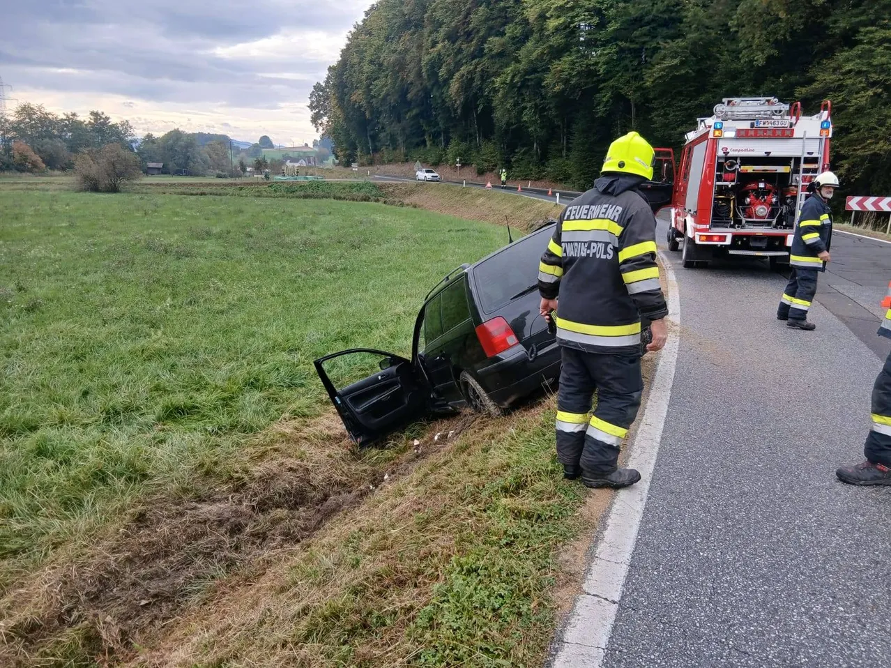 Ein Auto ist in einen Grasbereich neben der Straße gecrasht. Ein Feuerwehrmann steht neben dem Auto, und ein Feuerwehrwagen ist am Straßenrand geparkt.