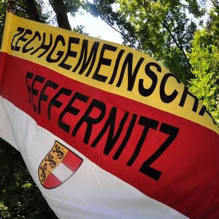 Ein Nahaufnahme eines Fahnens mit 'ZECHGEMEINSCHAFT SEFFERNITZ' in fetten schwarzen Buchstaben auf rotem und gelbem Hintergrund. Die Fahne zeigt ein Wappen mit einem roten und weißen Schild und drei goldenen Löwen.