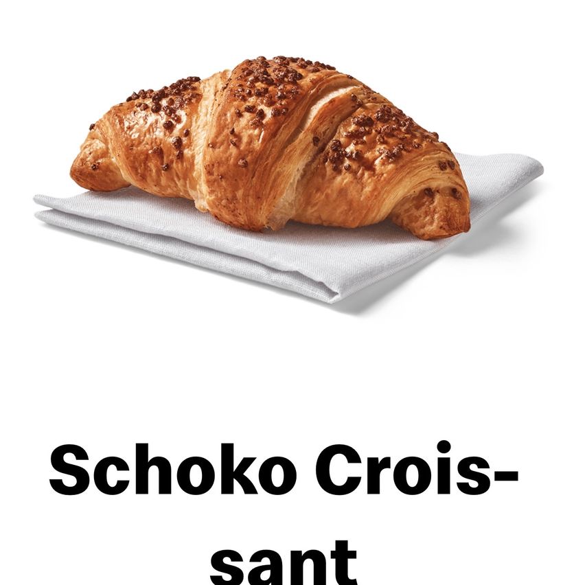 Ein mit Schokolade überzogenes Croissant mit goldener Kruste liegt auf einem weißen Serviettentuch. Darunter steht das Wort 'Schoko Crois-sant' in schwarzer Schrift.